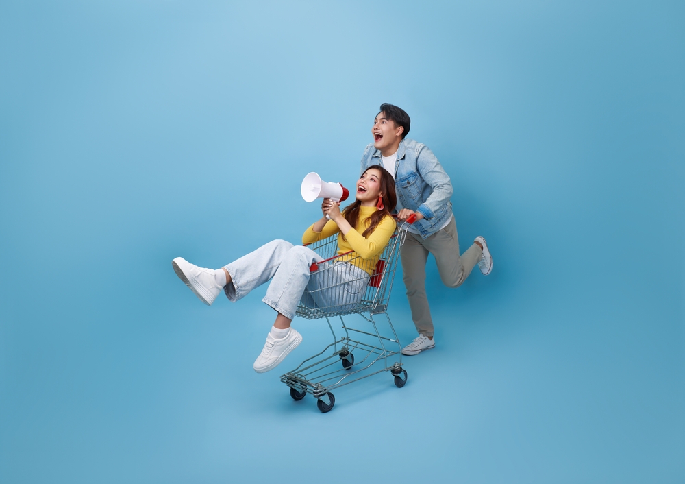 Young woman sitting in shopping trolley with a megaphone. She is being pushed by a man and they are both surprised and happy.